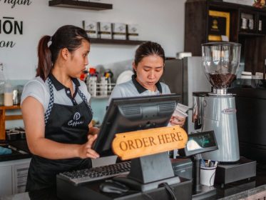 Two women at cafe cash register. Sign at register says "Order here"