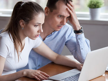 Man and woman sitting at a desk with an open laptop in front of them. They look stressed.