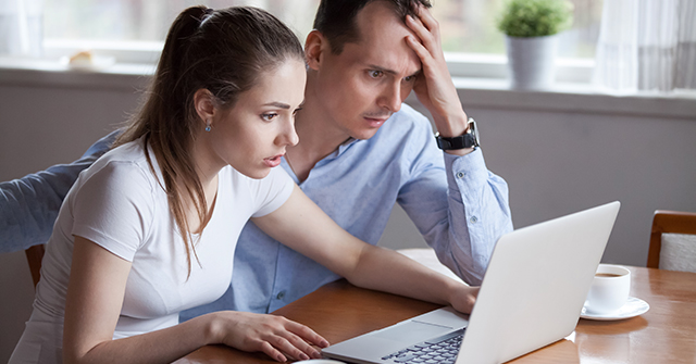 Man and woman sitting at a desk with an open laptop in front of them. They look stressed.