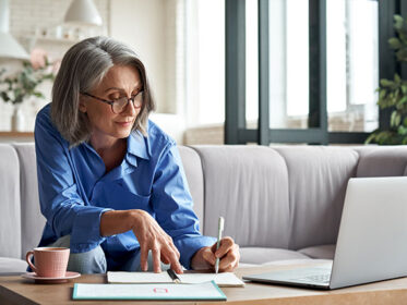 Mature lady sitting at a table in front of a laptop writing in a notebook