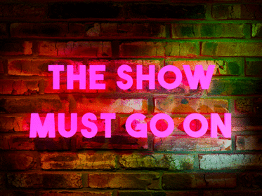 Image of a brick wall on which a pink neon sign reads, "The show much go on"