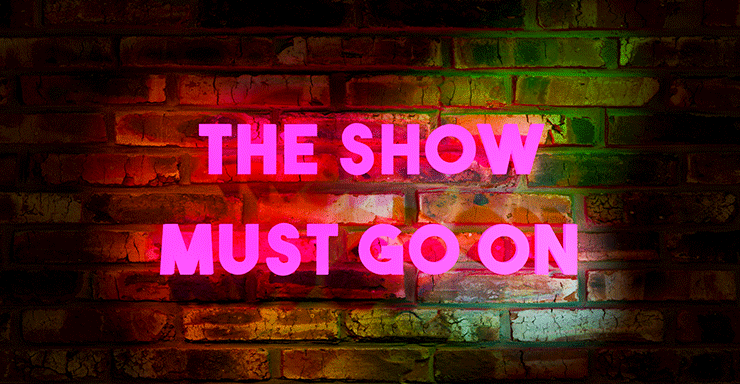 Image of a brick wall on which a pink neon sign reads, "The show much go on"