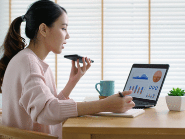Young asian female sits in a well lit room at her desk. She has her phone in her left hand, and laptop open infront of her. She speaks with an accountant on the phone as she analyses the graphs and statistics open on her computer screen. There is a blue mug sitting on the desk to her left, and a small luscious succulent to her right.