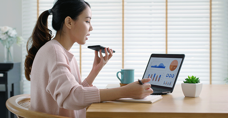 Young asian female sits in a well lit room at her desk. She has her phone in her left hand, and laptop open infront of her. She speaks with an accountant on the phone as she analyses the graphs and statistics open on her computer screen. There is a blue mug sitting on the desk to her left, and a small luscious succulent to her right.