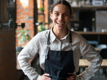 A young female cafe worker wearing a navy blue apron faces the camera with her hands on her hips with a big smile on her face.