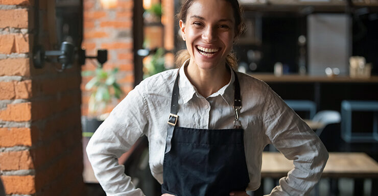 A young female cafe worker wearing a navy blue apron faces the camera with her hands on her hips with a big smile on her face.
