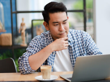 Asian Man in his mid 20s sits in a well lit room with his hand on his chin. He thoughtfully gazes at the laptop screen that is open in front of him.