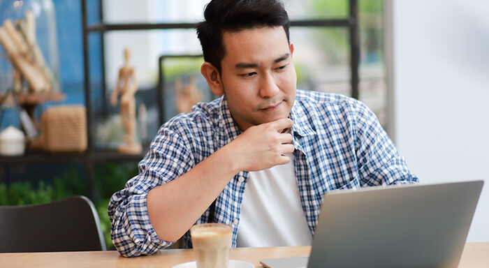 Asian Man in his mid 20s sits in a well lit room with his hand on his chin. He thoughtfully gazes at the laptop screen that is open in front of him.